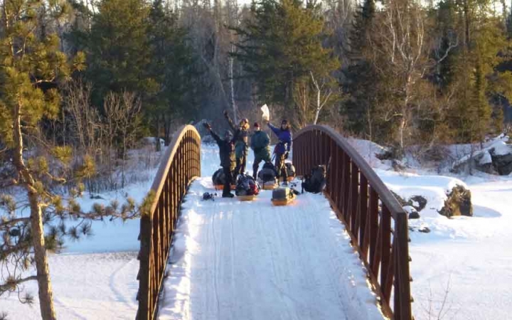 a group of cross country skiers, each pulling small sleds, make their way across a snowy bridge